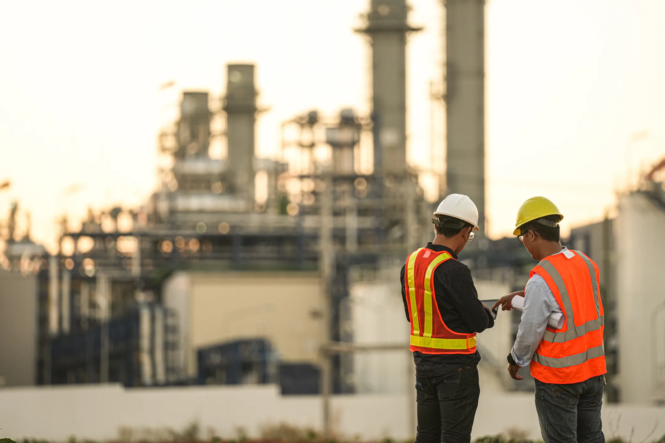 Two people with hard hats and safety vests with their backs to the camera stand in front of a facility.