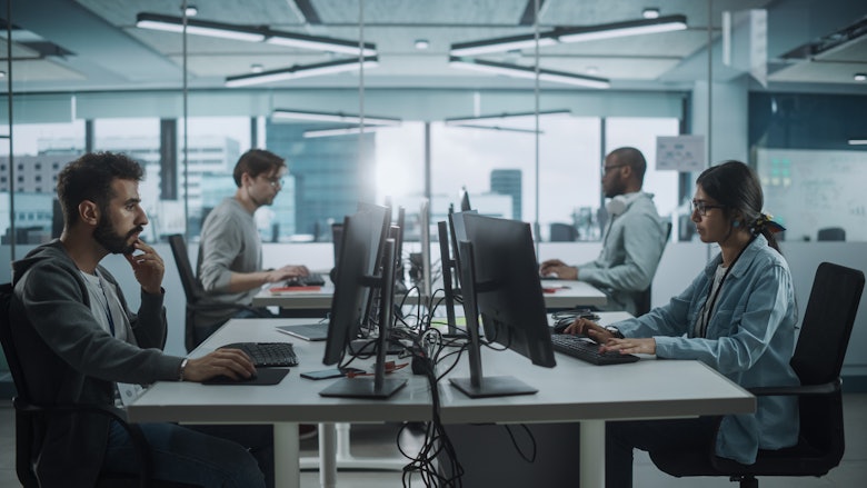 Workers sit at tables working on computers in an open-concept office.