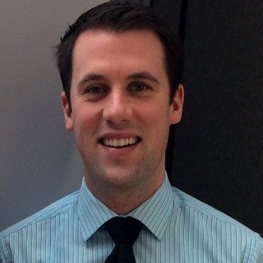 A headshot of a smiling dark-haired man in a blue-striped shirt and dark tie.