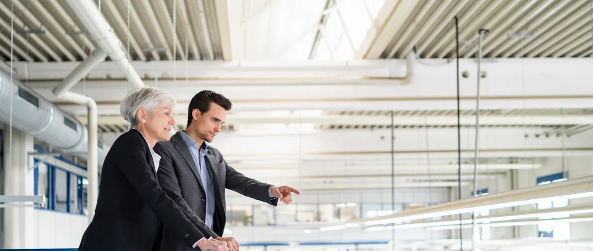 A woman with short gray hair wearing a black pantsuit stands on a walkway overlooking a factory floor, next to a younger man with dark hair and wearing a gray suit.