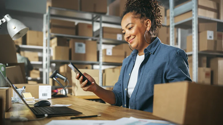 A woman with dark curly hair wearing an open blue button-down shirt with a white t-shirt underneath is seen smiling as she looks at her smartphone in a storage warehouse.