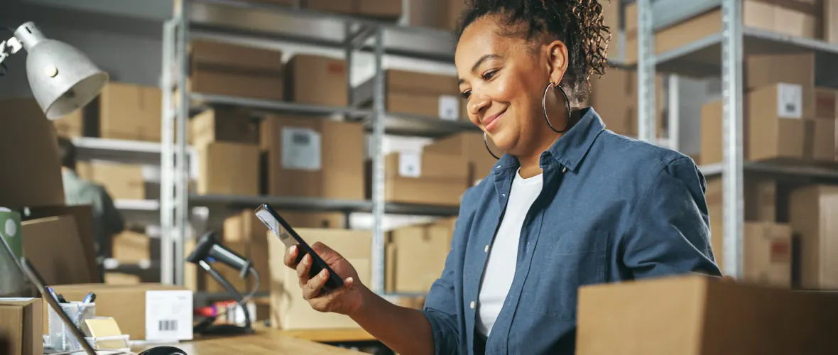 A woman with dark curly hair wearing an open blue button-down shirt with a white t-shirt underneath is seen smiling as she looks at her smartphone in a storage warehouse.