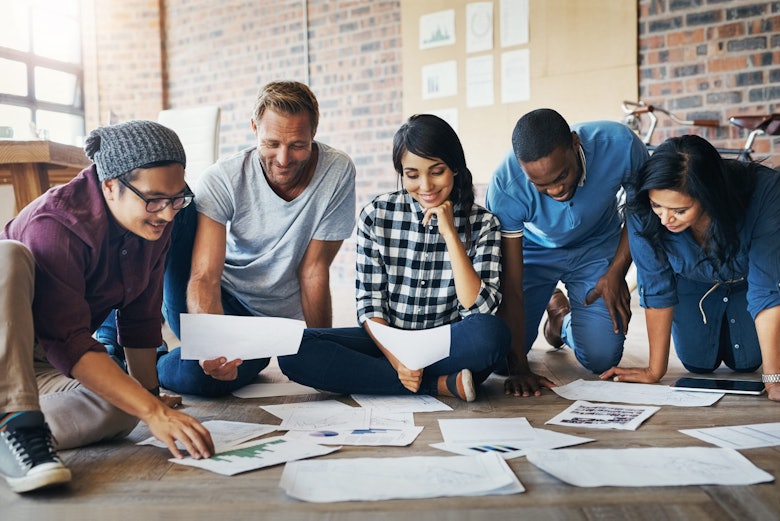 Five people in casual clothes sit on the floor of an office, sorting through papers.