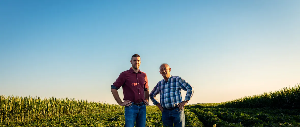 A younger man in a red shirt and an older man in a blue and white shirt stand in a field at dusk.