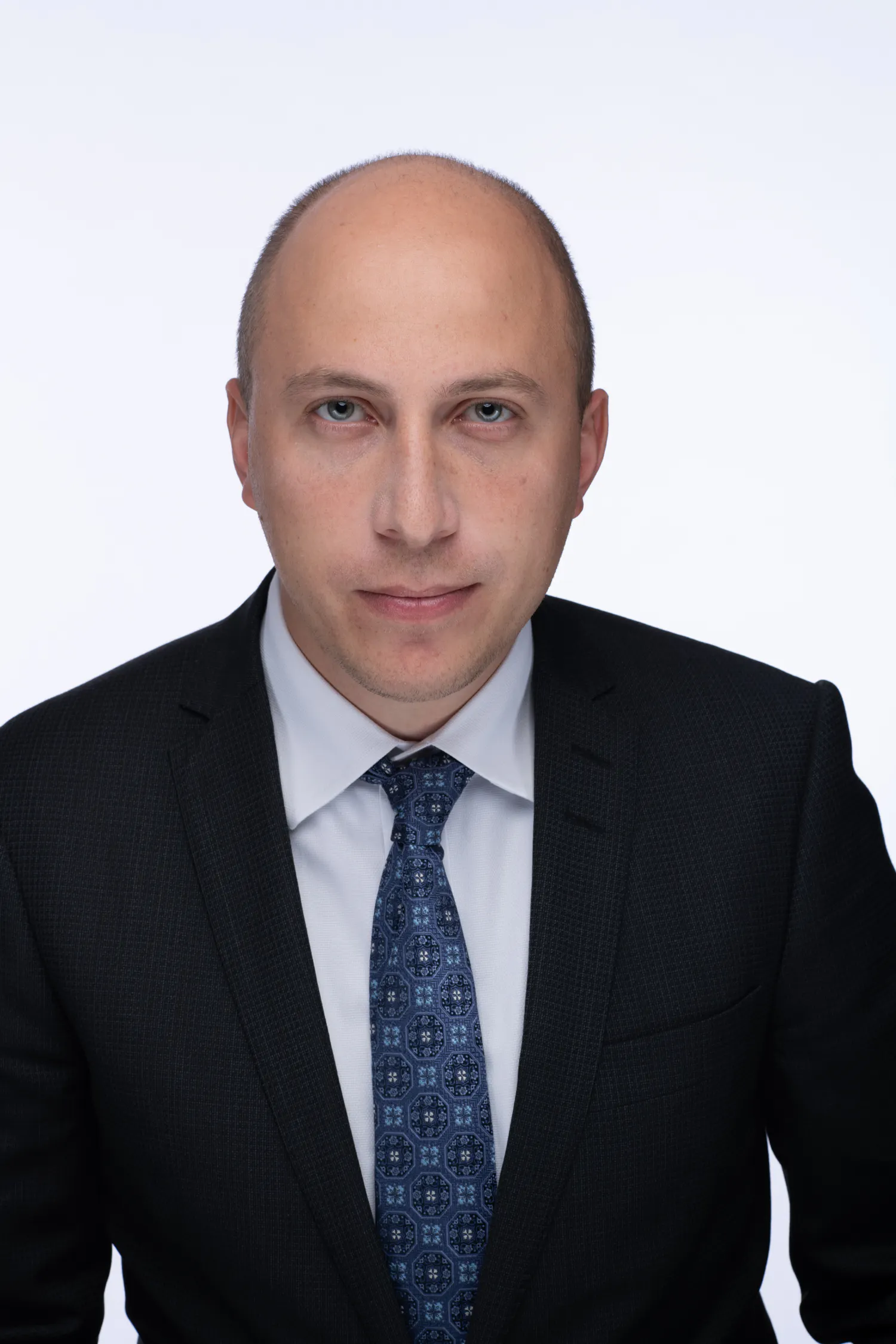 A man in a black suit and blue-patterned tie looks straight at the camera.