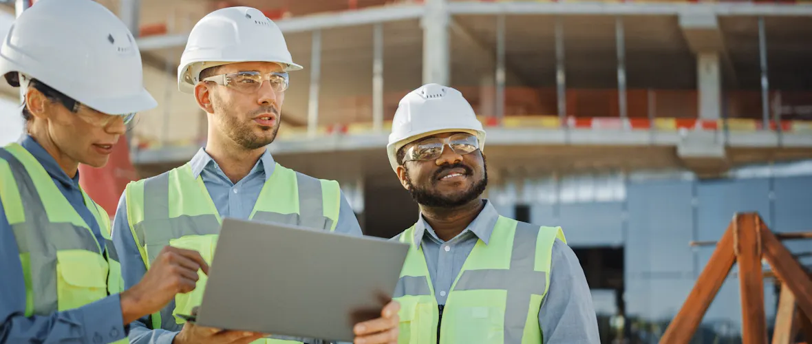 Three people wearing white hard hats and yellow safety vests look at a tablet at a construction site.