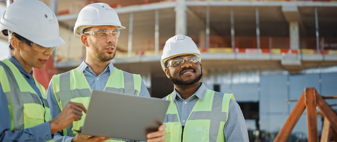 Three people wearing white hard hats and yellow safety vests look at a tablet at a construction site.