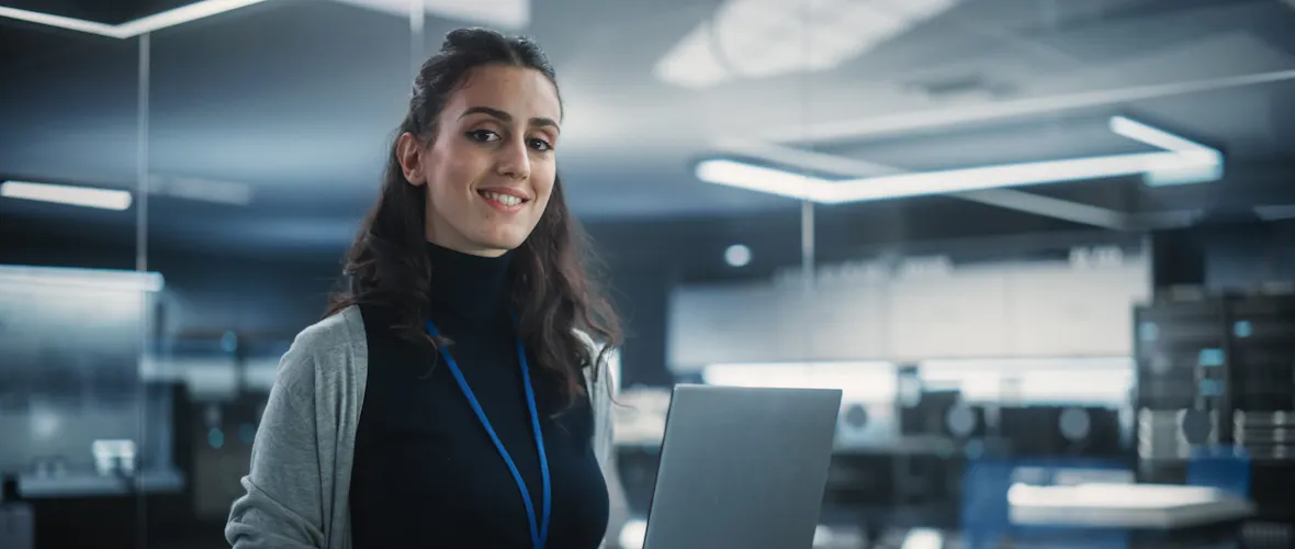 A young woman with dark hair partly tied back, wearing a black top under a grey cardigan, hold a laptop and smiles at the camera.