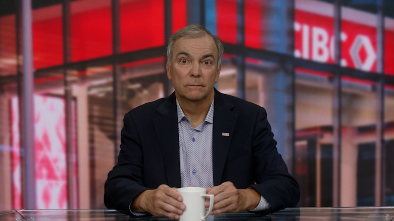 A man wearing a dark jacket and light blue shirt sits behind a desk with a white coffee mug in front of him.