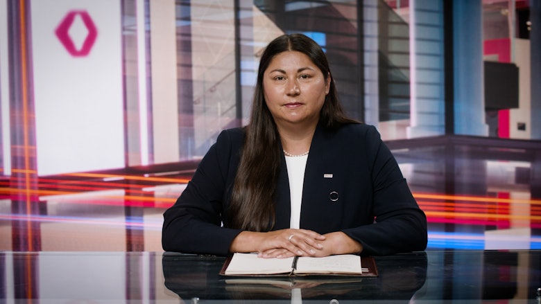 A woman with long dark hair, wearing a white shirt and black blazer, sits behind a desk and smiles at the camera.