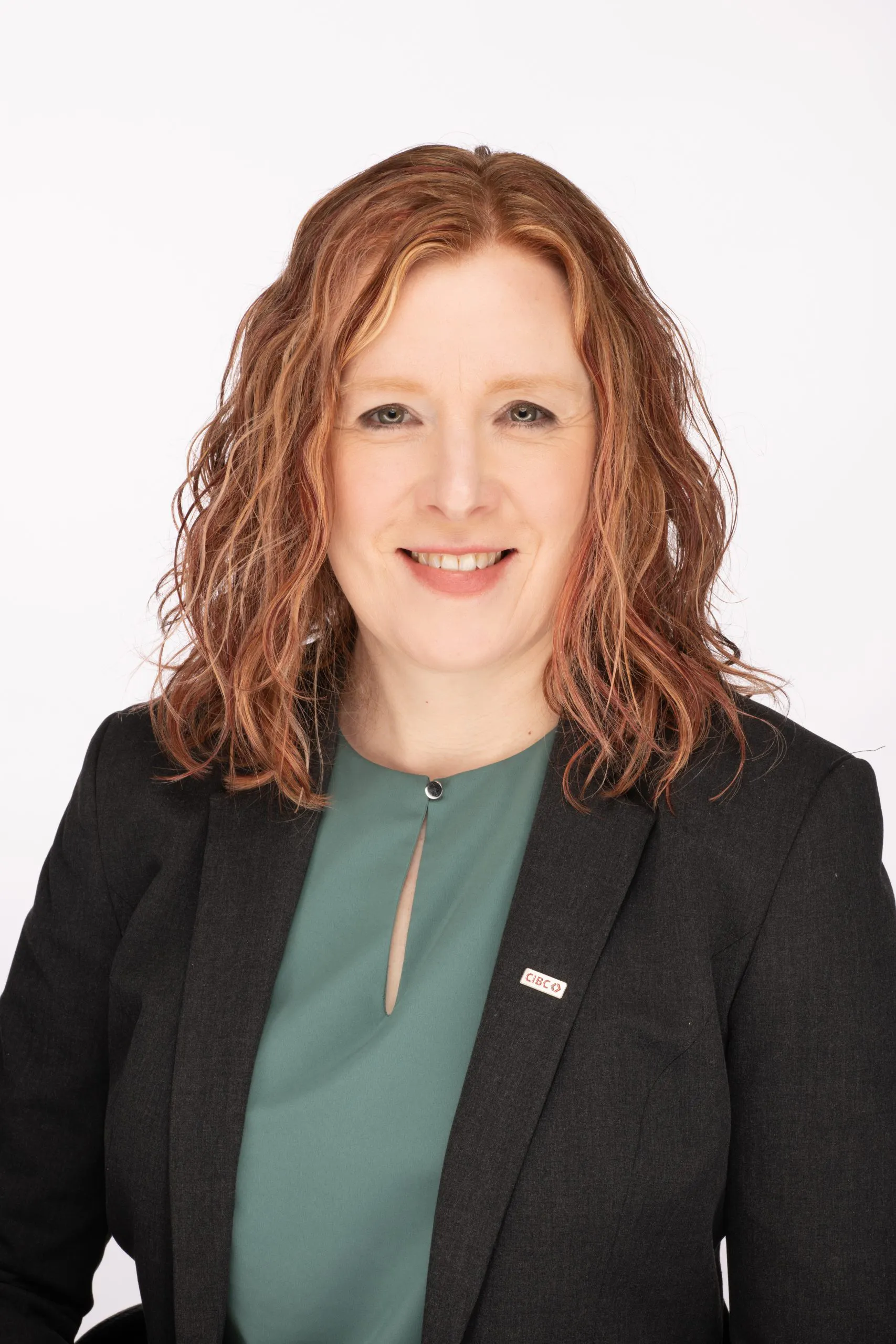 A woman with curly red hair wearing a black blazer and a green shirt smiles at the camera.