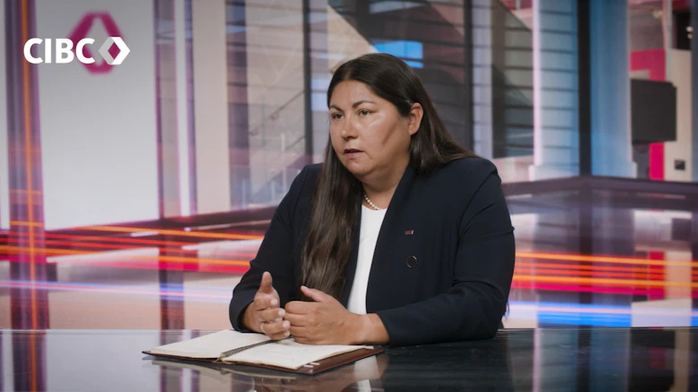 Une femme aux longs cheveux foncés, portant une chemise blanche sous un blazer noir, est assise à un bureau et regarde hors caméra.