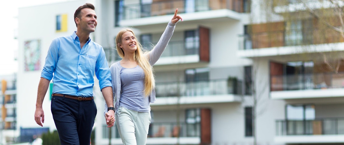 A man in a light blue shirt and dark blue pants holds hands with a woman with long blonde hair, wearing a pale lavender twinset, khakis and high heels smile as they walk around an apartment complex.