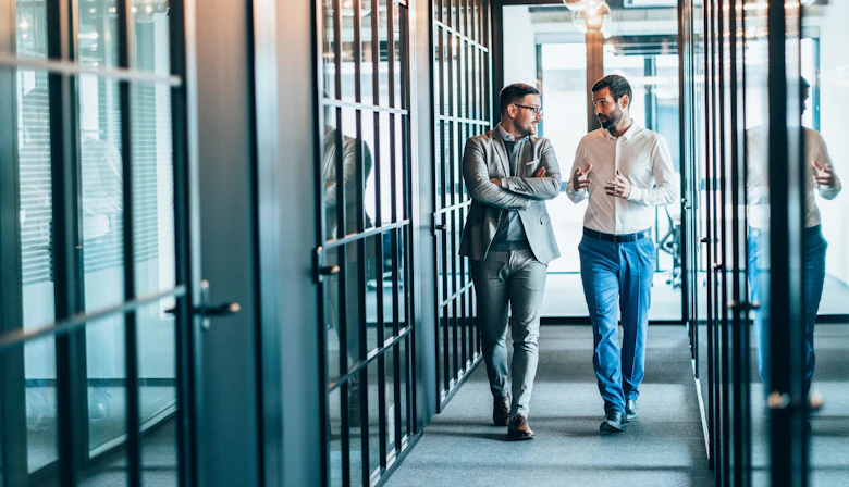 Two men walk down a hallway lined with glass-doored offices while looking at each other and talking.