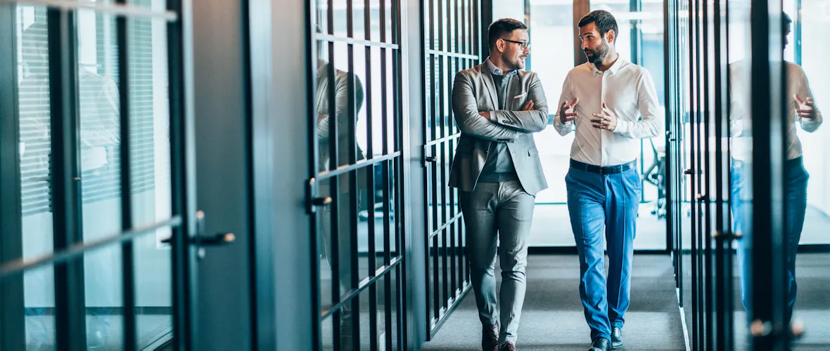 Two men walk down a hallway lined with glass-doored offices while looking at each other and talking.