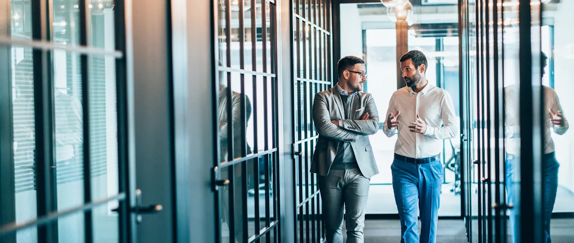 Two men walk down a hallway lined with glass-doored offices while looking at each other and talking.