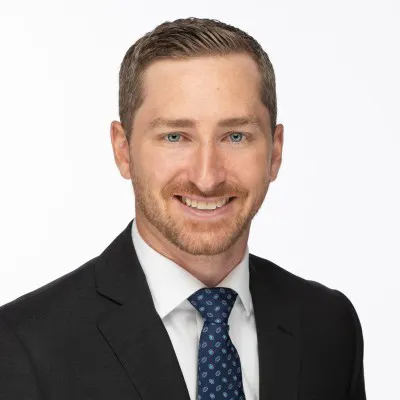 A man with close-cropped blond hair and beard and wearing a dark suit, white suit and blue tie, smiles for the camera.
