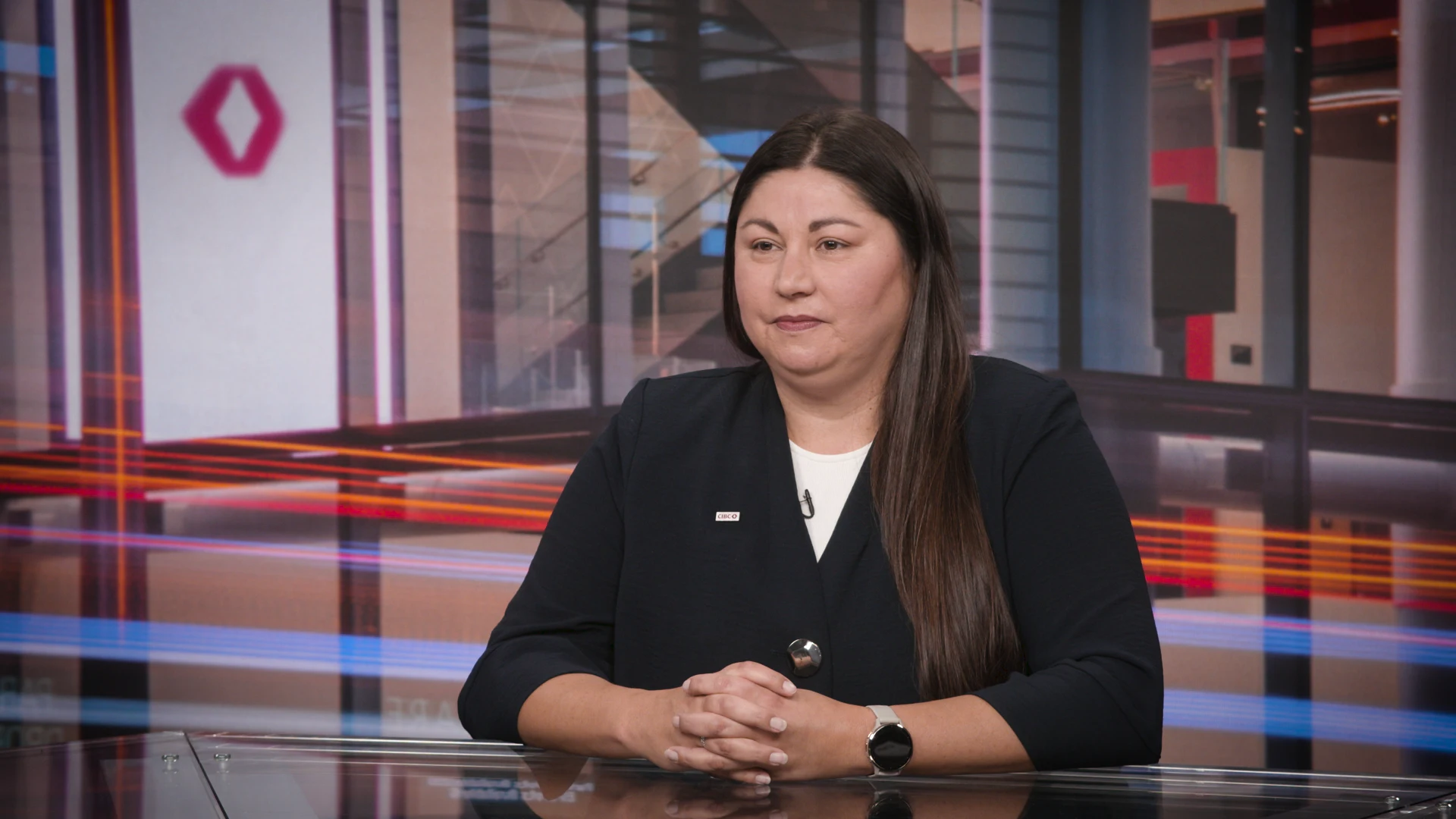Une femme aux longs cheveux foncés, portant un blazer noir sur une chemise blanche, est assise à un bureau, les mains croisées.