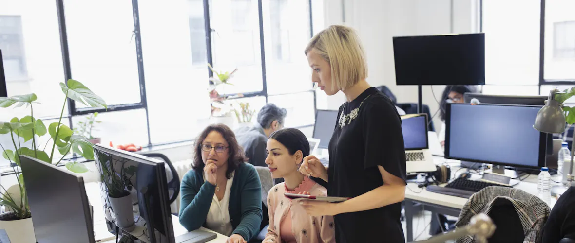 Three women - two seated and one standing - gather around a desk to look at a computer screen.