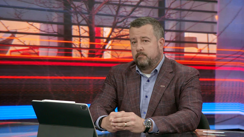 Un homme avec une barbe, portant une veste mauve sur une chemise bleue, est assis à un bureau, les mains croisées.
