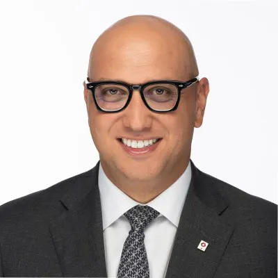 A man wearing dark-framed glasses, a dark suit, a white shirt and a patterned tie, smiles at the camera.