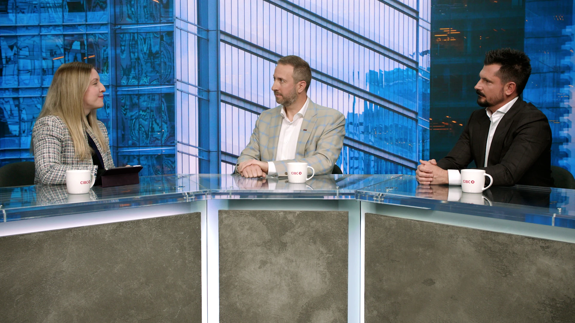 Two men sit behind a desk, with hands folded, as they look at a woman who is also sitting at the desk, with an iPad in front of her.