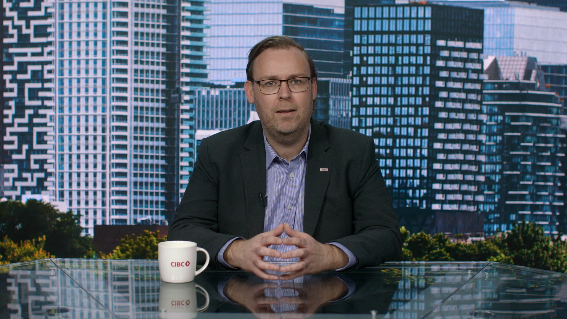 A man wearing glasses, a dark jacket and a blue shirt sits behind a desk and looks at the camera.