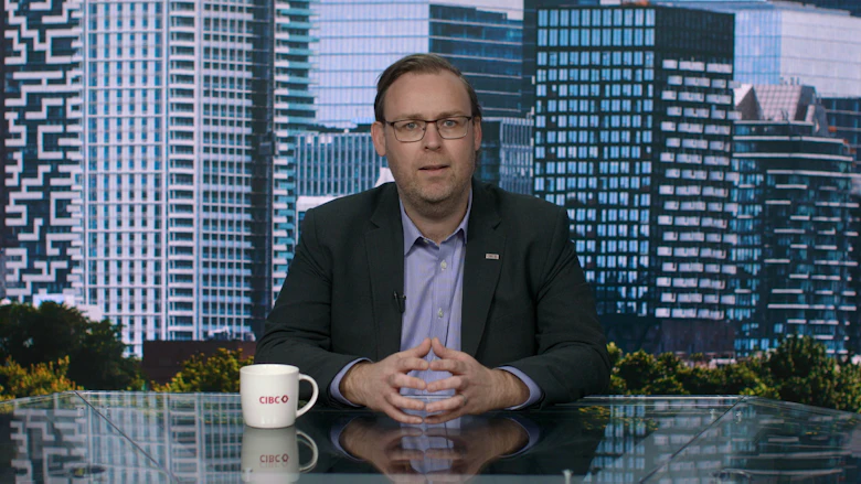 A man wearing glasses, a dark jacket and a blue shirt sits behind a desk and looks at the camera.