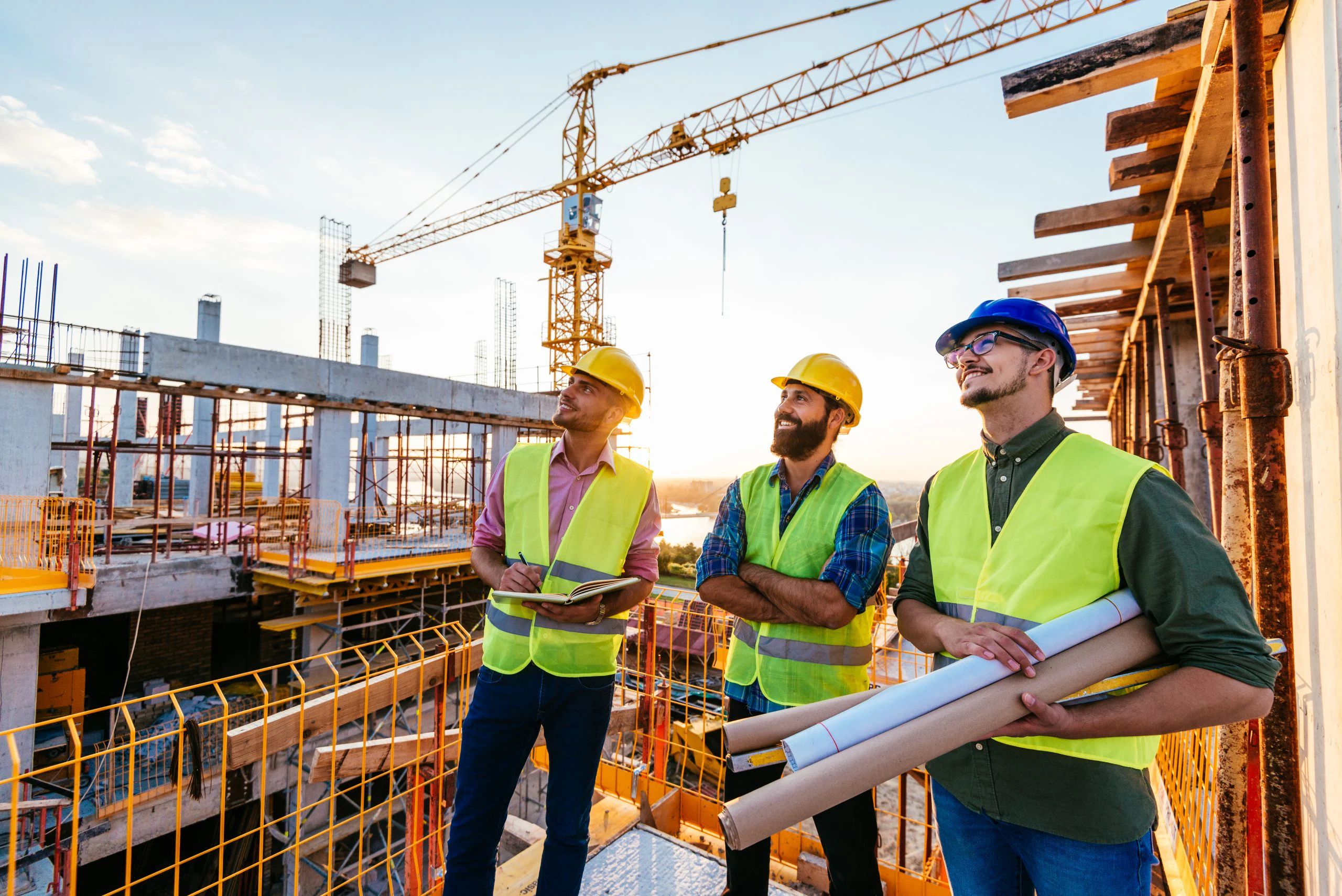Trois hommes portant des casques de chantier et des gilets de sécurité, debout sur une plateforme sur un grand chantier de construction, regardent vers la gauche en souriant.