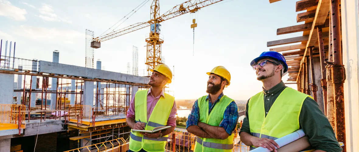 Three men wearing construction hates and safety vests standing on a platform at a large building construction site look off to the left, smiling.