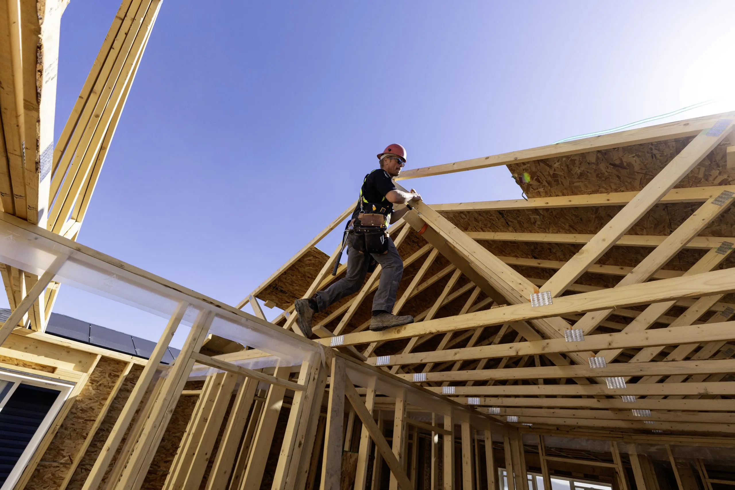 Un ouvrier du bâtiment travaille sur l’ossature en bois d’une maison sous un ciel bleu.
