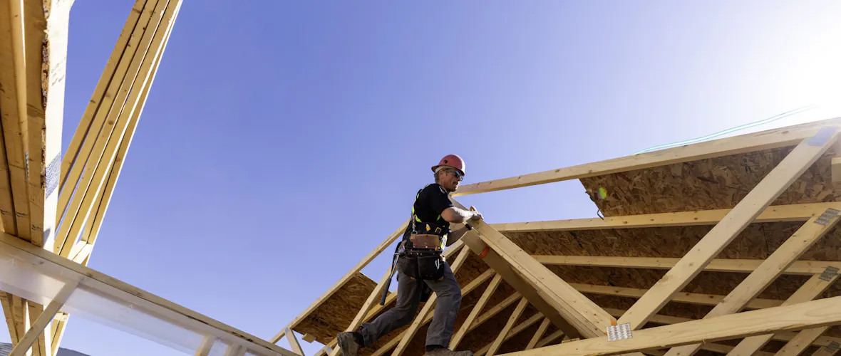 A construction worker works on the wooden frame of a house under a blue sky.