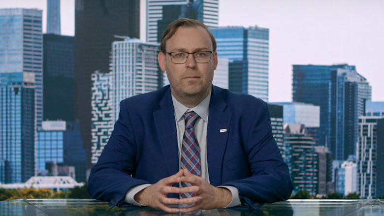 A man in a medium-blue suit and a light grey shirt and checked tie sits with his hands folded and looks at the camera.