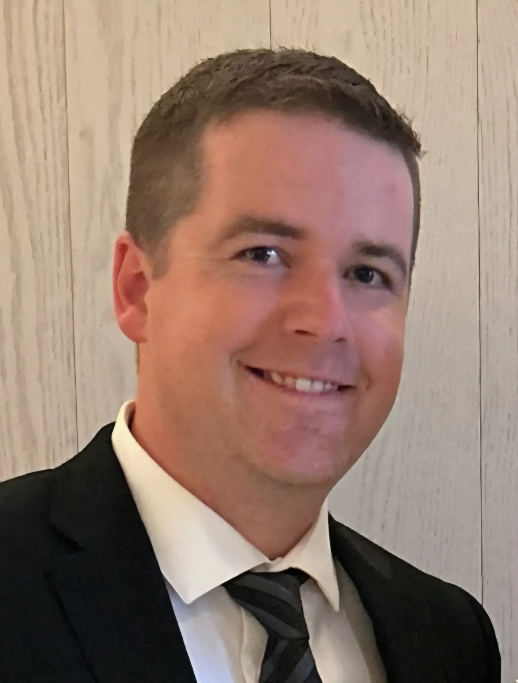 A man with light brown hair, wearing a dark suit jacket, white shirt and black tie, smiles for the camera.