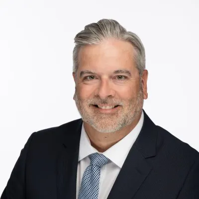 A man with grey hair and a close-shaven grey beard, wearing a dark suit jacket, white shirt and light blue tie, smiles at the camera.
