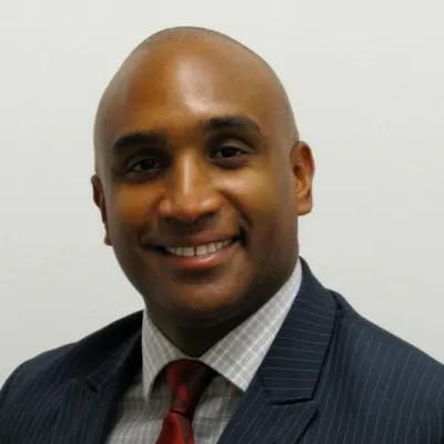 A man with a clean-shaven head, wearing a dark suit jacket and a red tie, smiles at the camera.