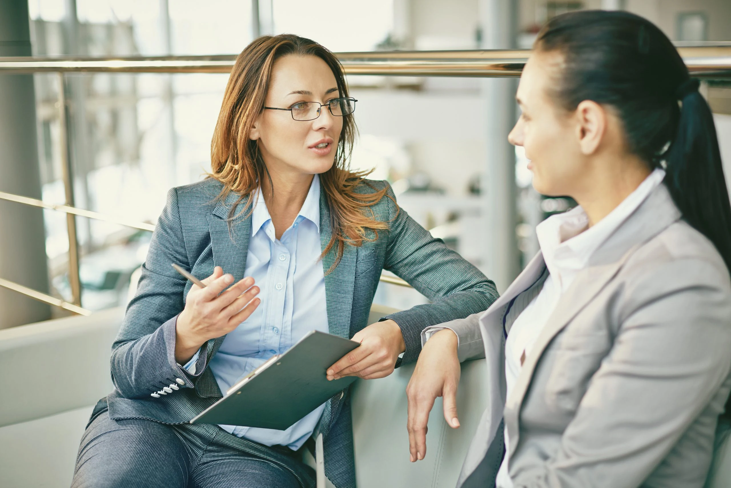 A woman with layered, light-brown hair wearing glasses, a light grey suit and a light blue shirt, holds a pen and clipboard while talking to a colleague with dark hair wearing a white shirt and light jacket.