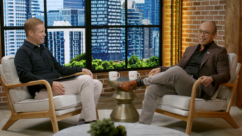 Two men in dark suit jackets and light-coloured pants sit facing each other in white armchairs with Toronto's skyline through a window behind them.