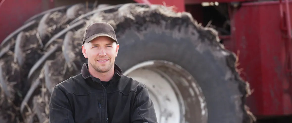 Un homme portant un pull sombre et une casquette grise se tient devant un grand tracteur, les bras croisés, souriant à la caméra.