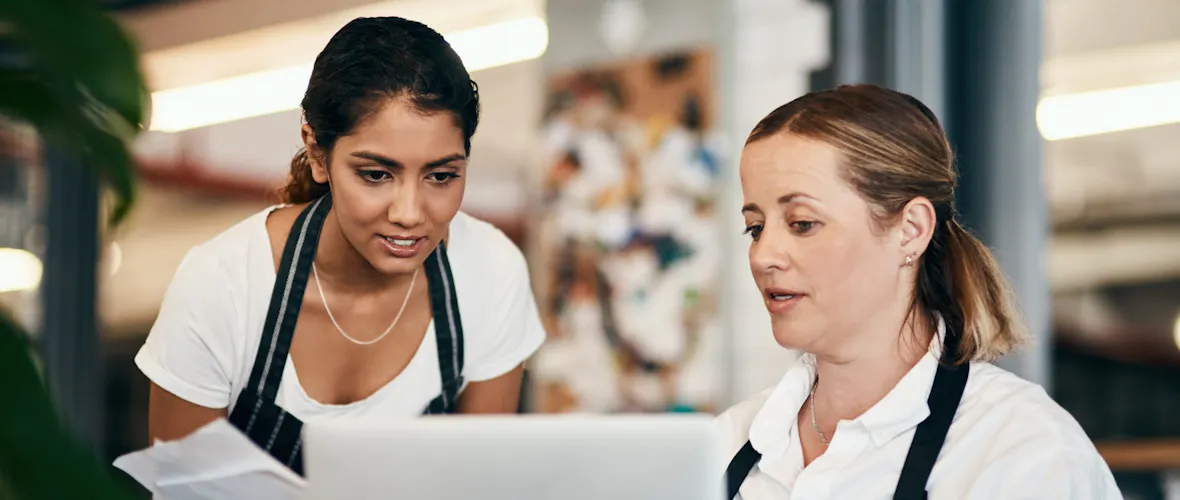 Two women - one with blond hair tied back and another with brown hair tied back - both wearing white shirts and dark aprons, consult a laptop.